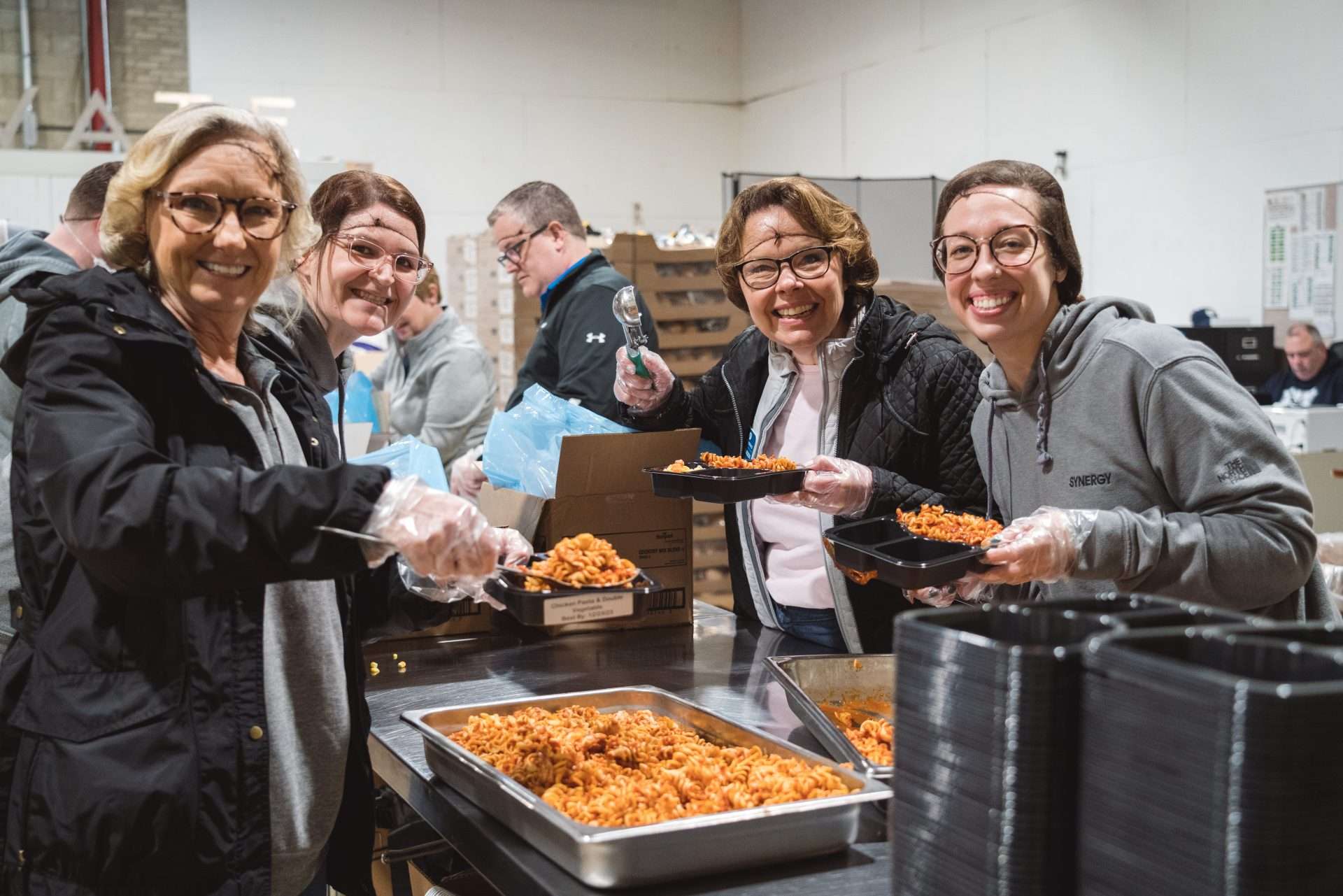 Four women working in a commercial food rescue kitchen serving up pasta and smiling at the camera
